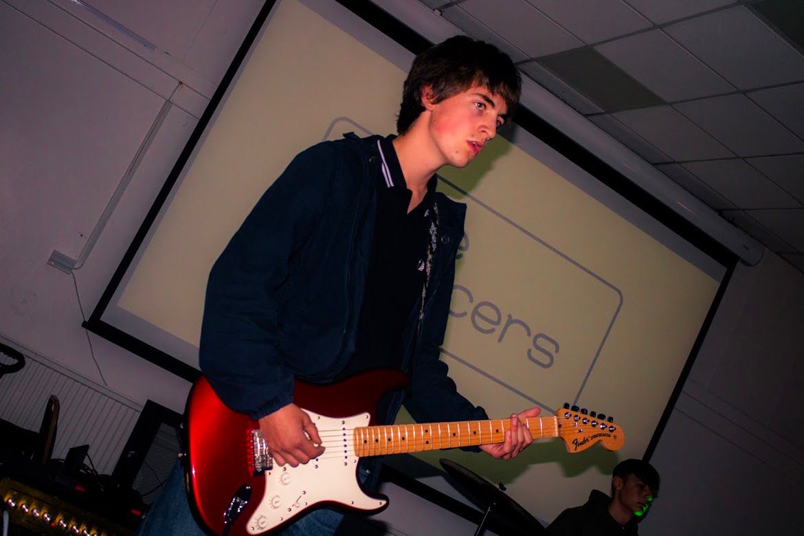 Harry Kelly of The Fleecers playing a red Fender Stratocaster in front of the band's projected logo